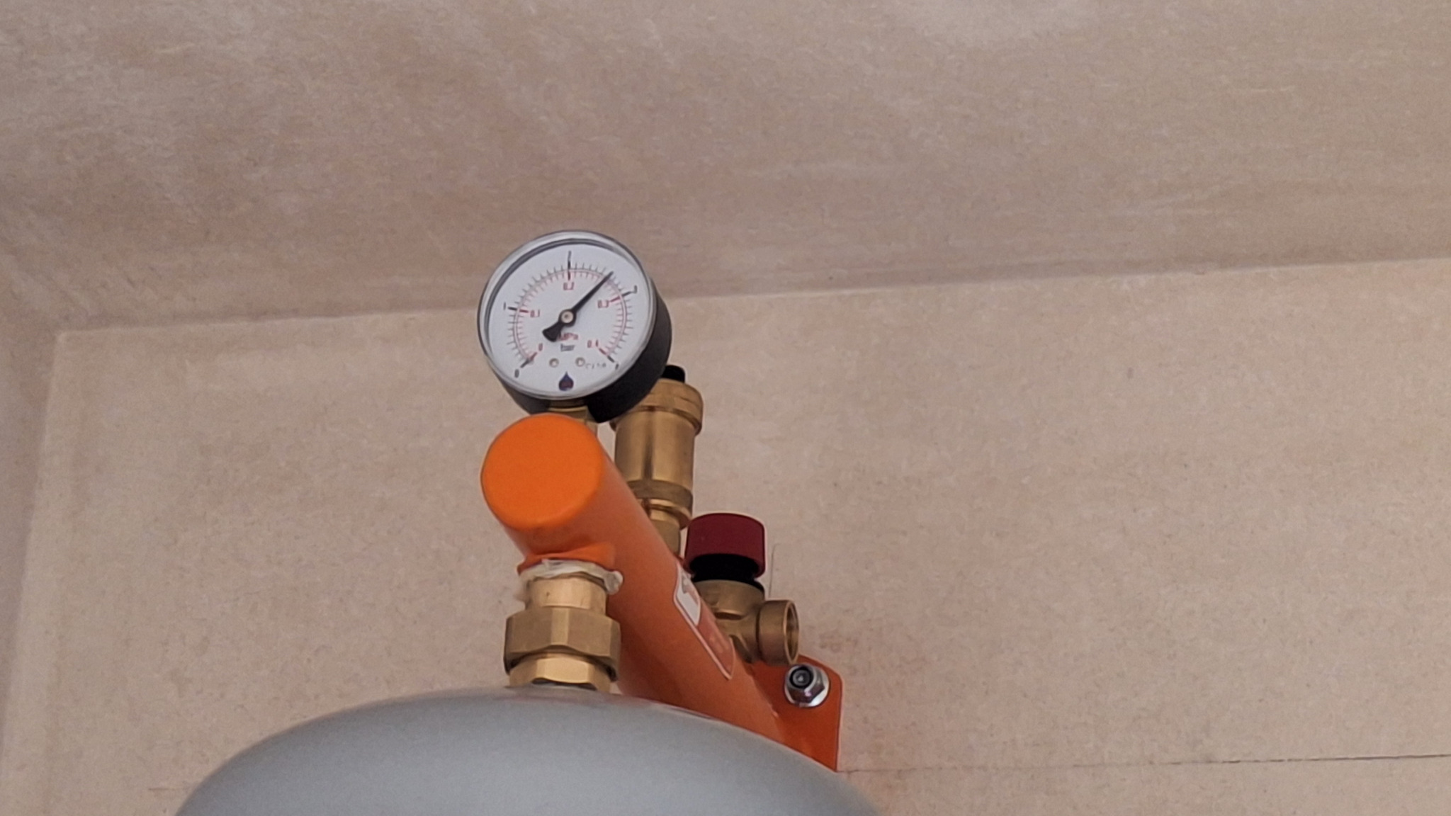 Manometr na pompie ciepła z oranżową rurą i czerwonym zaworem, widoczne przyłącza mosiężne oraz fragment szarego zbiornika.