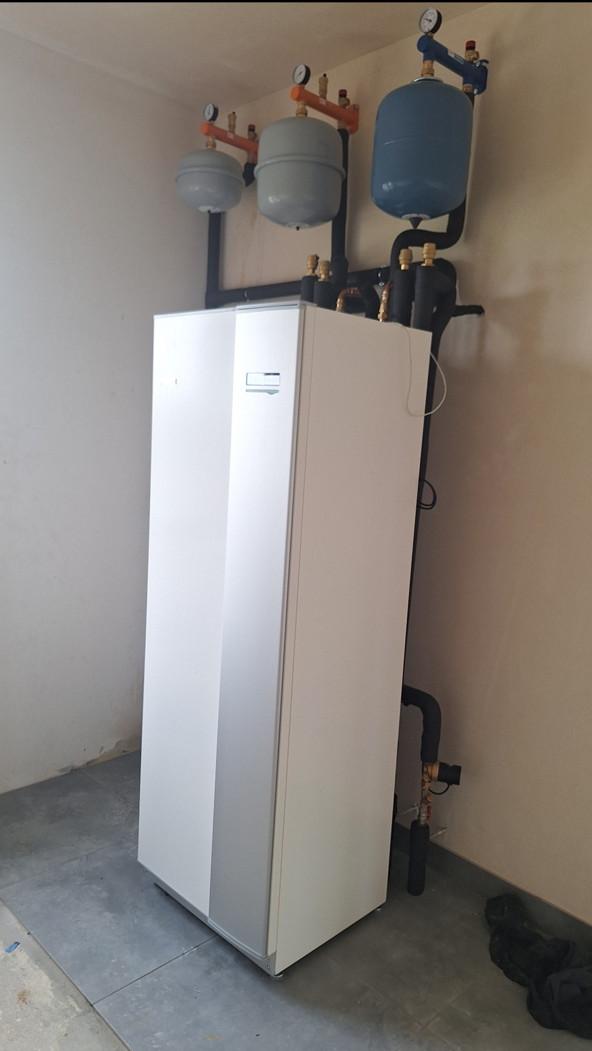 Biała jednostka wewnętrzna z szarym panelem, z trzema zbiornikami wyrównawczymi na górze, zamontowana w rogu pomieszczenia z surowymi ścianami.