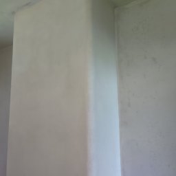 Dariusz Ostasiewicz - Świeżo otynkowany narożnik w pomieszczeniu, widoczne ślady po tynkowaniu na ścianie w tle. Jasne, naturalne światło podkreśla gładką powierzchnię tynku.