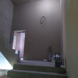 Dariusz Ostasiewicz - Wnętrze w trakcie prac wykończeniowych: otynkowane ściany, schody w surowym stanie, widoczne przewody i narzędzia. Oświetlenie żarówką na kablu.