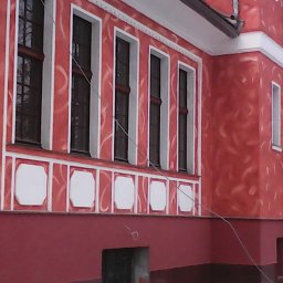 Dariusz Ostasiewicz - Czerwona fasada budynku z dekoracyjnymi tynkami i białymi obramowaniami okien. Widoczne detale architektoniczne i wzory na ścianie.