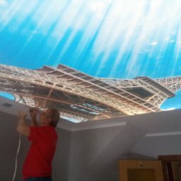 Dariusz Ostasiewicz - Montaż sufitu napinanego z motywem wieży Eiffla. Instalator w czerwonej koszulce mocuje konstrukcję. Widok z dołu na sufit i fragment ściany.