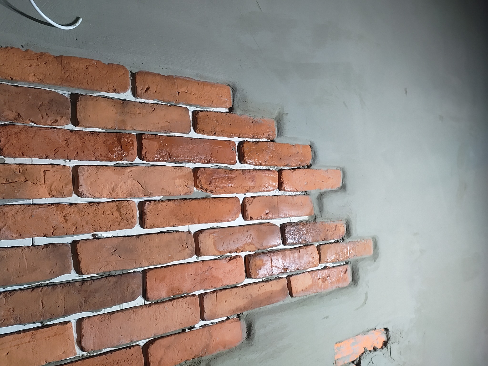 Fragment ściany z dekoracyjną cegłą w trakcie montażu. Widoczne świeże zaprawy i niedokończone krawędzie. Oświetlenie podkreśla fakturę cegieł.