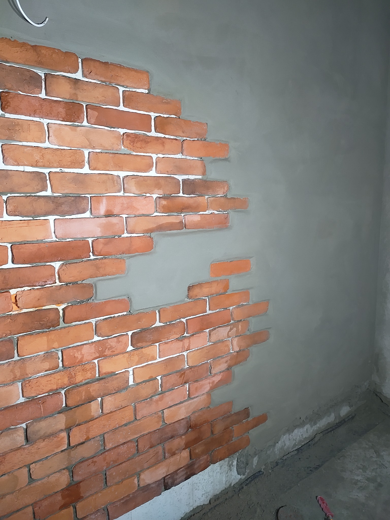 Ściana z częściowo ułożoną dekoracyjną cegłą, kontrastująca z surowym tynkiem. Widoczne niedokończone prace, podkreślające etap realizacji.