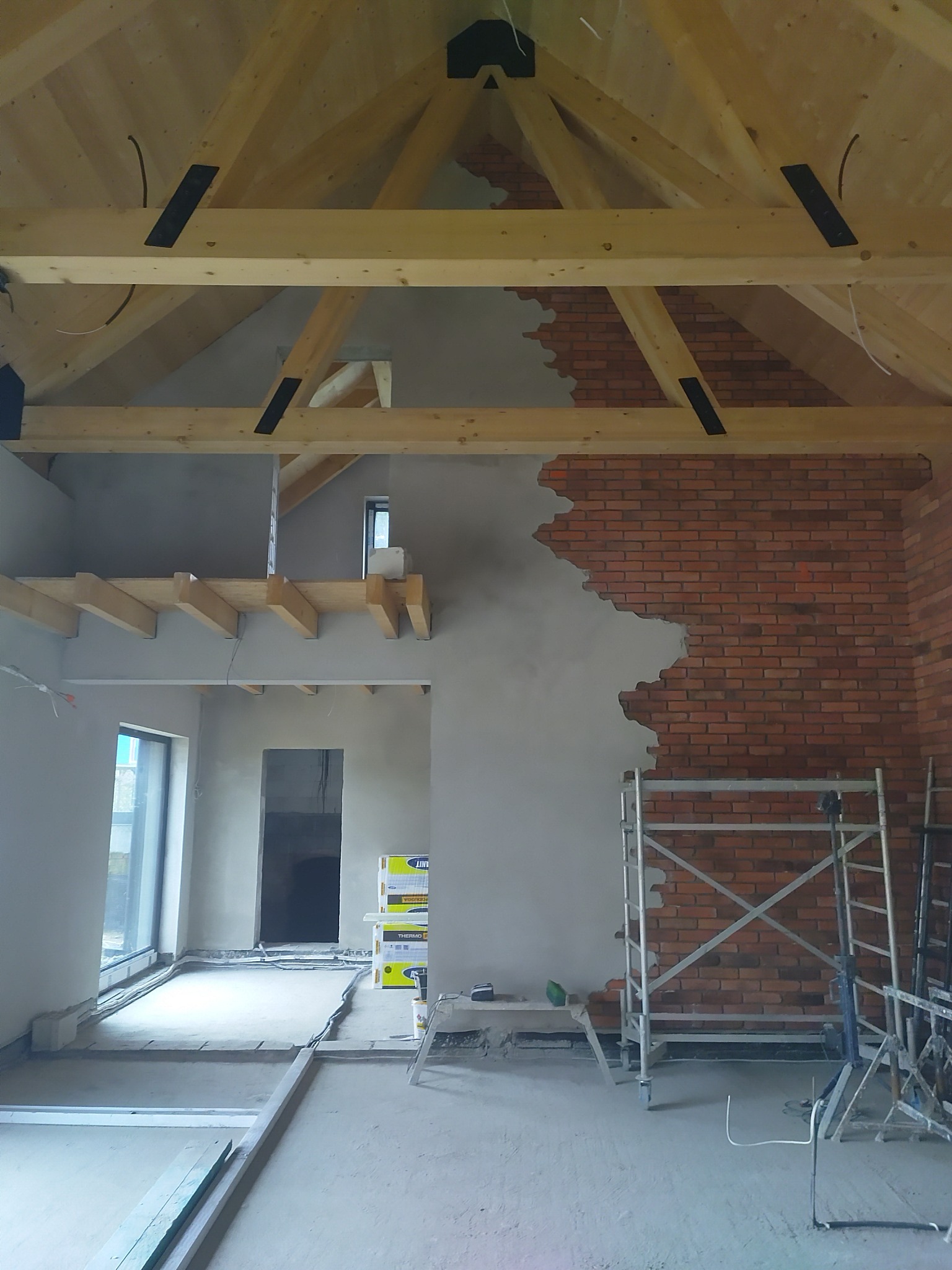 Surowy stan wnętrza z drewnianą konstrukcją dachu, częściowo otynkowaną ścianą z ceglanym wykończeniem i rusztowaniem. Widoczne przygotowania do dalszych prac wykończeniowych.