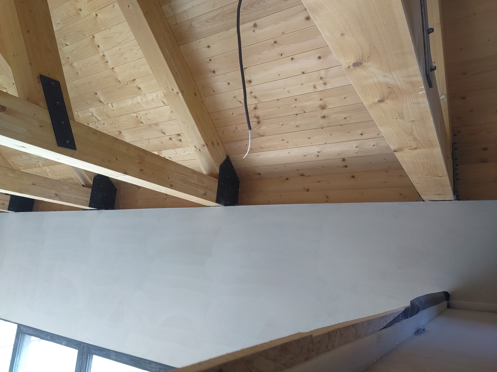 Drewniany strop z widocznymi belkami i metalowymi łącznikami, świeżo otynkowana ściana, fragment okna w dolnym rogu. Widok z dołu, perspektywa z narożnika pomieszczenia.