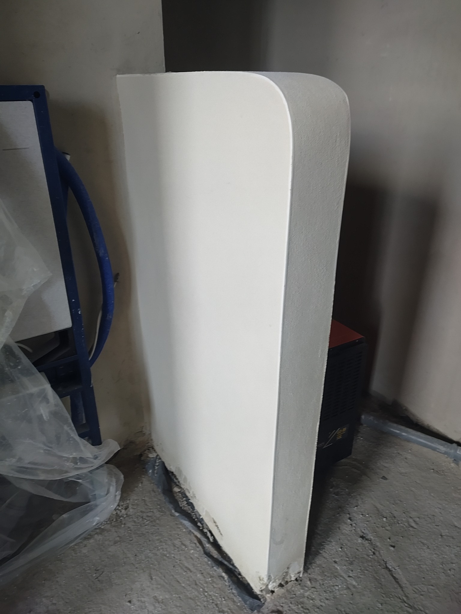 Biała, zaokrąglona obudowa stelaża WC w surowym stanie, przed wykończeniem. Widoczne elementy instalacji i podłoże w trakcie prac. Ujęcie z bliska, z boku.