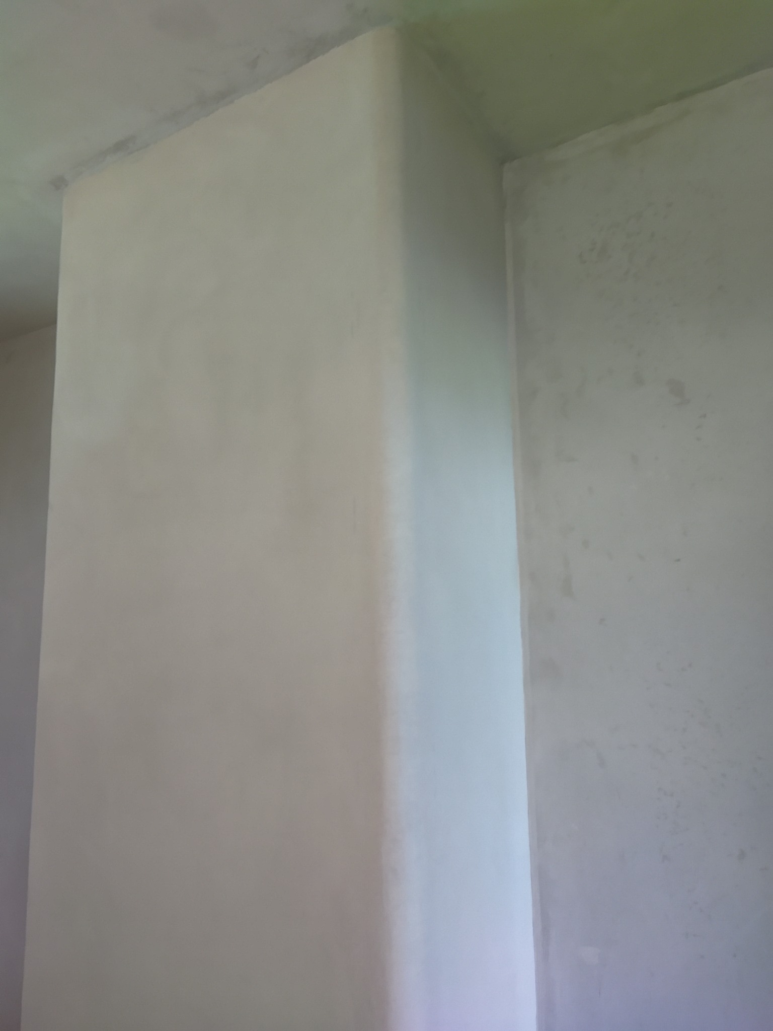 Świeżo otynkowany narożnik w pomieszczeniu, widoczne ślady po tynkowaniu na ścianie w tle. Jasne, naturalne światło podkreśla gładką powierzchnię tynku.