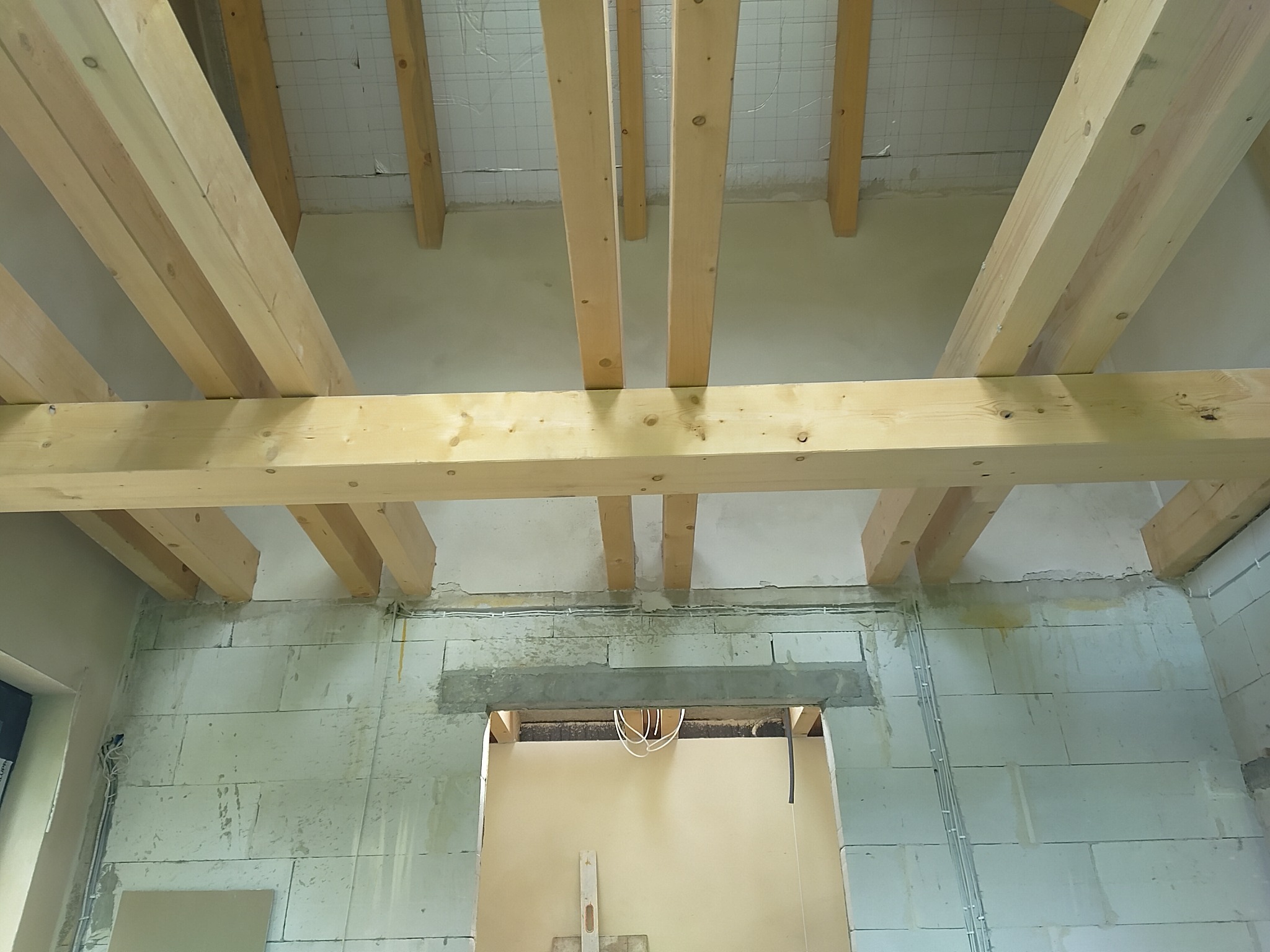 Surowy stan wnętrza z widoczną konstrukcją drewnianą stropu i ścianami z betonu komórkowego. Widoczne przewody elektryczne i otwór drzwiowy.