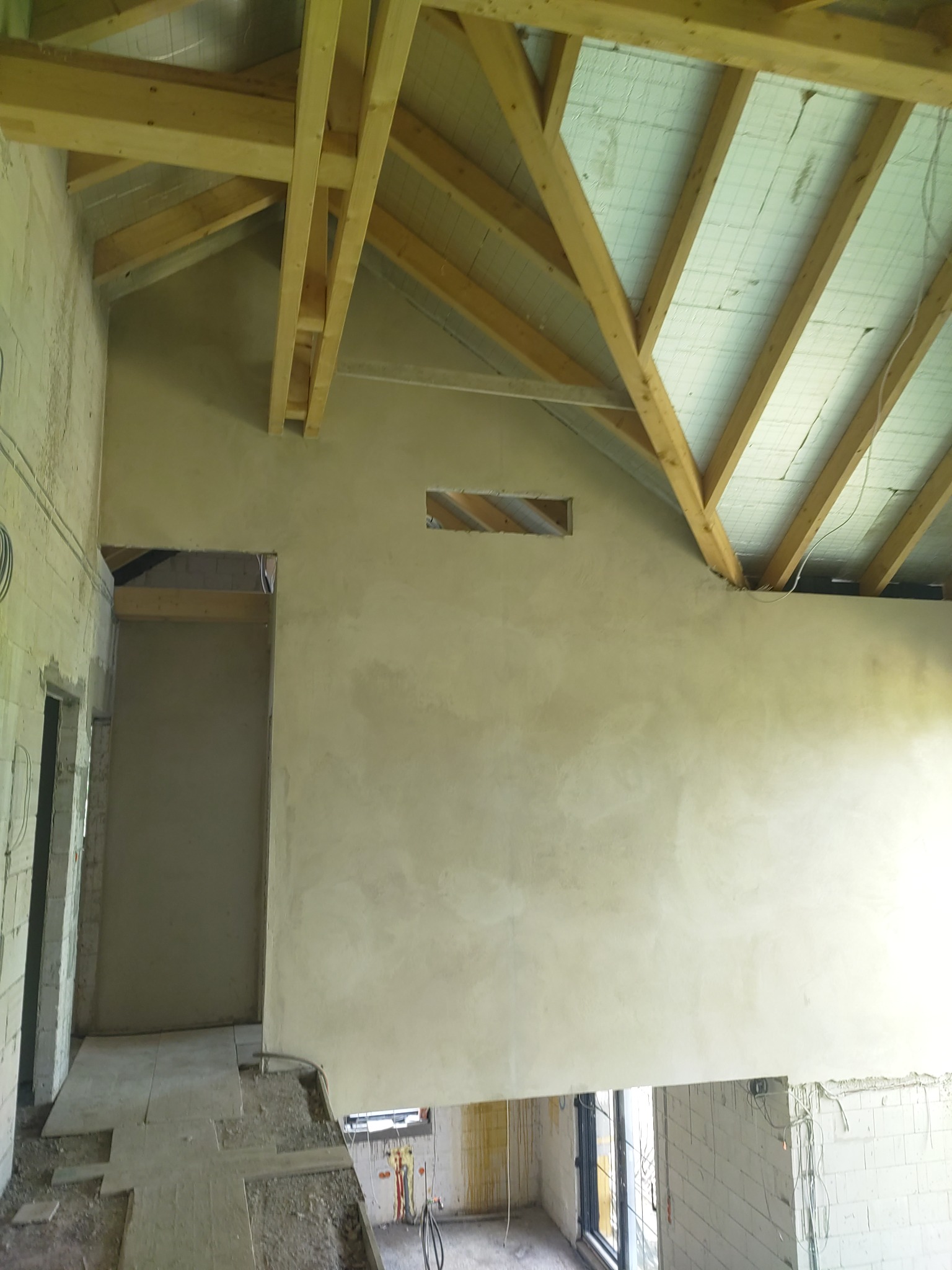 Wnętrze w trakcie tynkowania: surowa ściana z widoczną konstrukcją drewnianą dachu i oknem. Widoczne przewody elektryczne i fragment podłogi w trakcie prac wykończeniowych.
