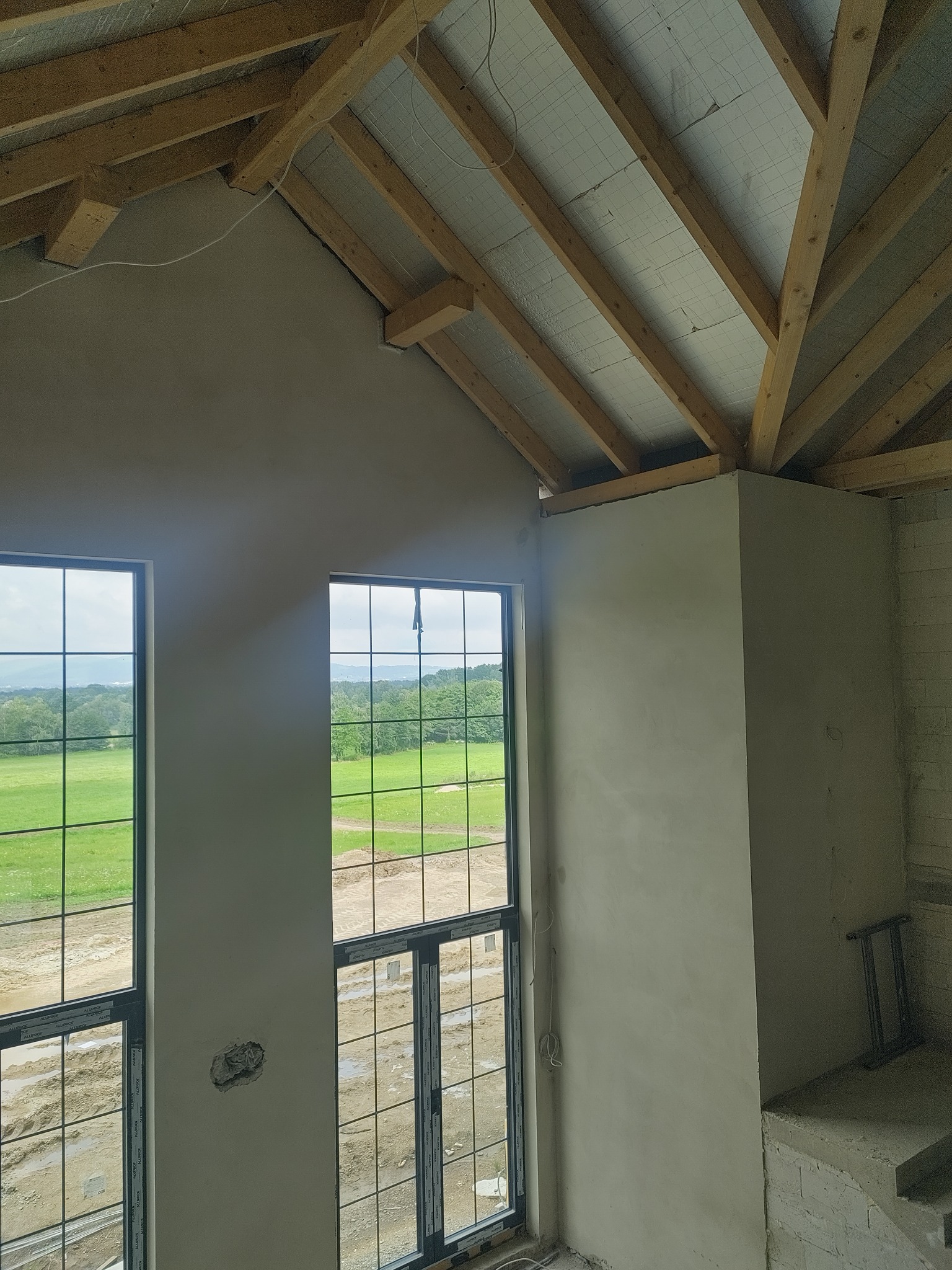 Wnętrze w stanie deweloperskim: świeżo otynkowane ściany, duże okna z widokiem na zieleń, drewniana konstrukcja dachu. Widoczny fragment drabiny.