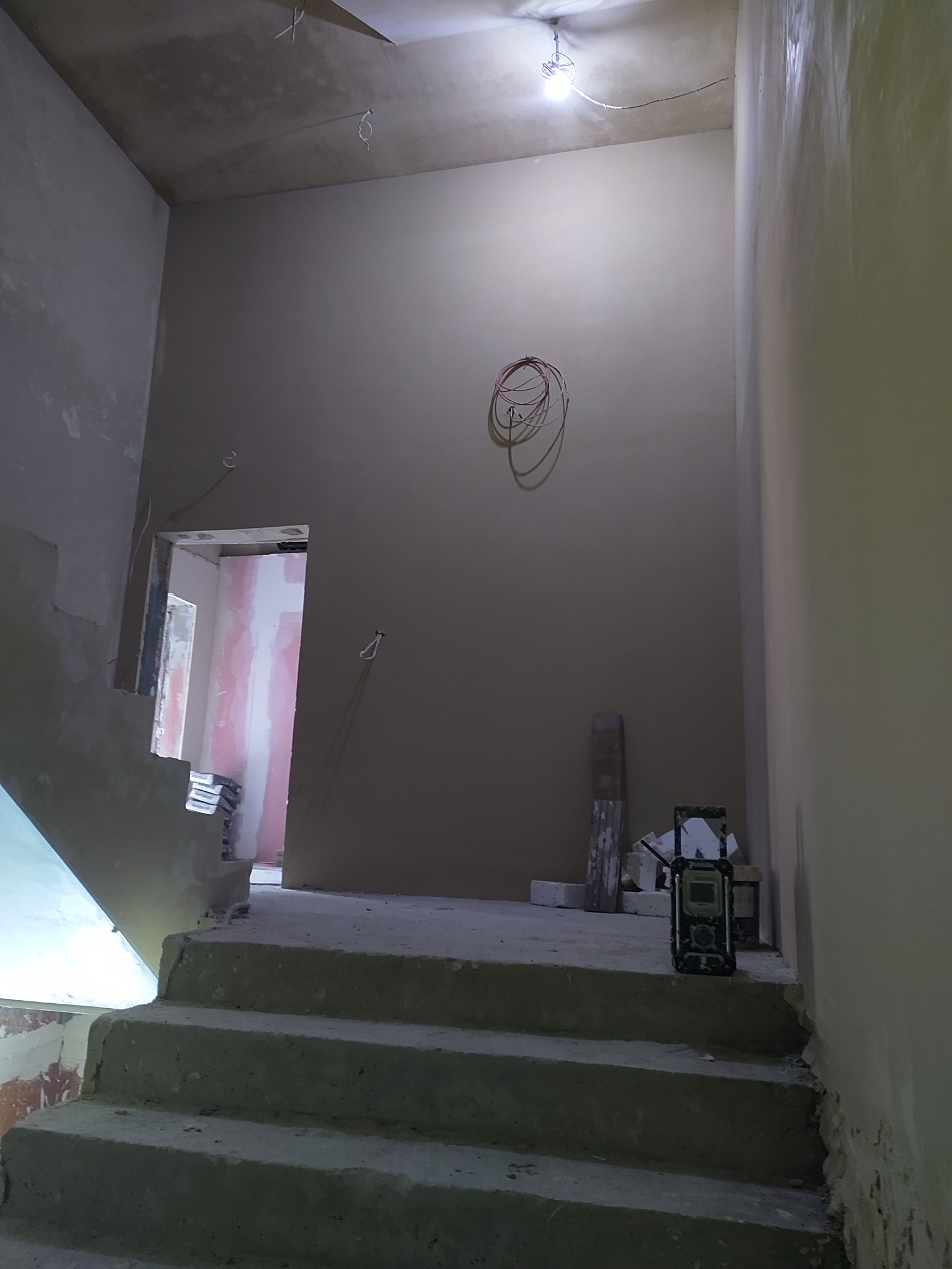 Wnętrze w trakcie prac wykończeniowych: otynkowane ściany, schody w surowym stanie, widoczne przewody i narzędzia. Oświetlenie żarówką na kablu.