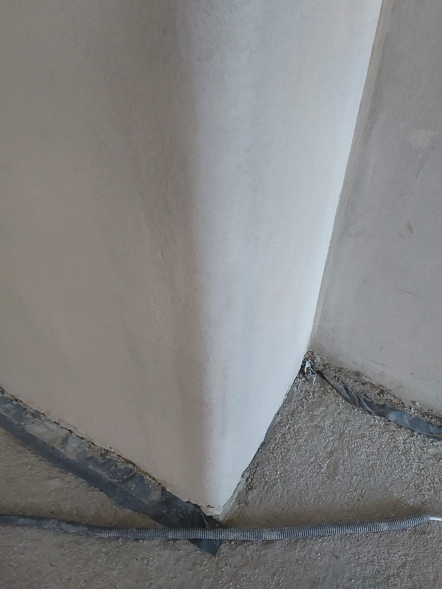 Świeżo otynkowany narożnik ścian w jasnym odcieniu, z widocznymi elementami zabezpieczającymi podłogę i kablem w peszlu przy podłodze. Widok z dołu.