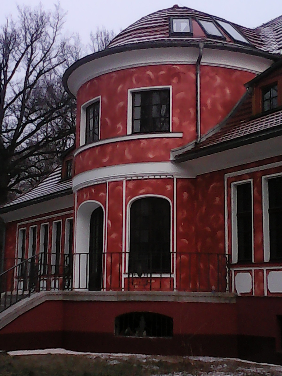 Czerwony budynek z dekoracyjnym tynkiem i białymi detalami architektonicznymi, widoczny fragment balkonu z czarną balustradą i okna.
