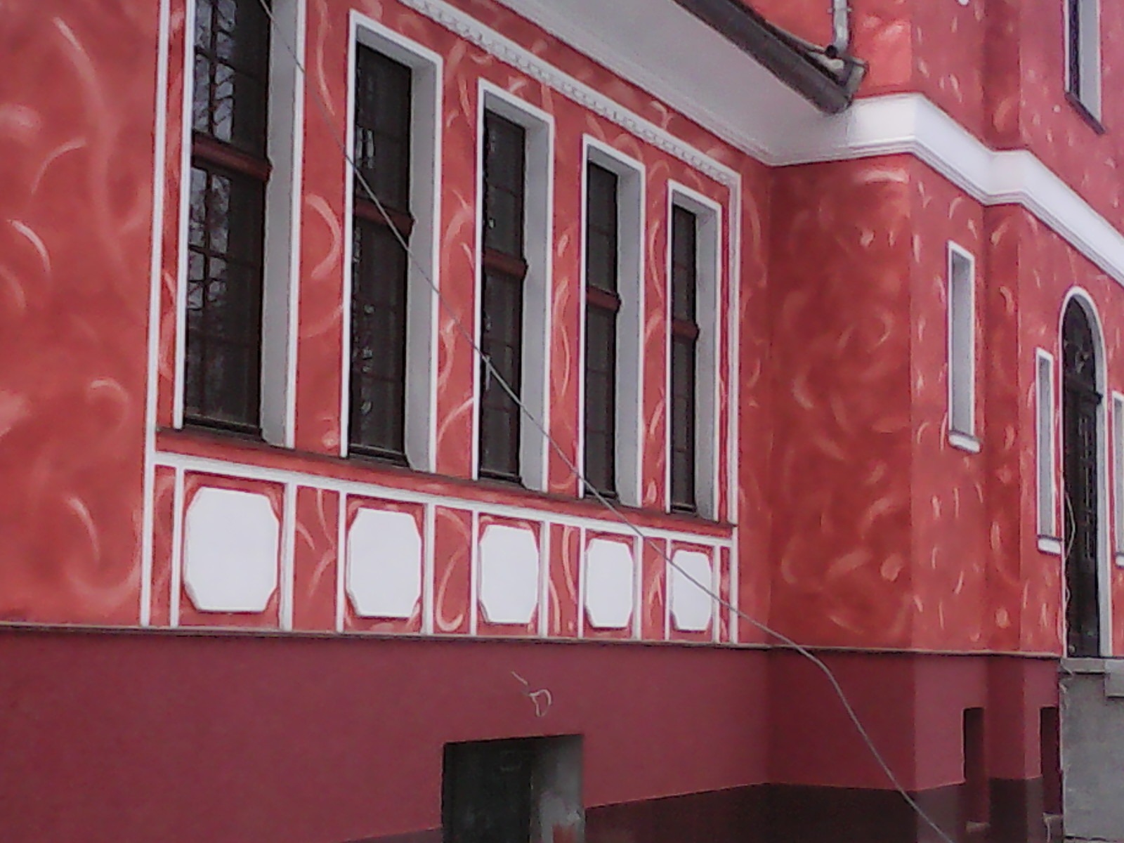 Czerwona fasada budynku z dekoracyjnymi tynkami i białymi obramowaniami okien. Widoczne detale architektoniczne i wzory na ścianie.