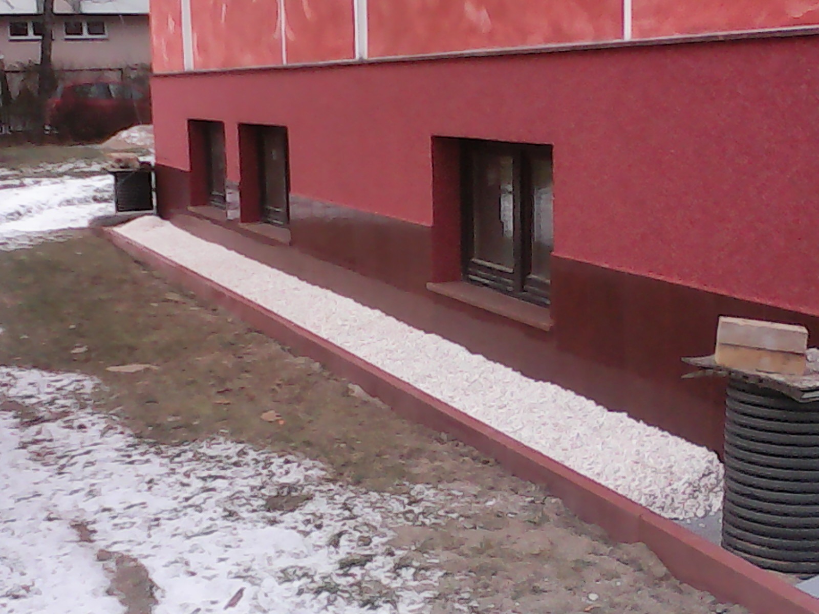Czerwona elewacja budynku z trzema oknami, obok drenaż z białego żwiru i rura spustowa. Częściowo ośnieżony teren.