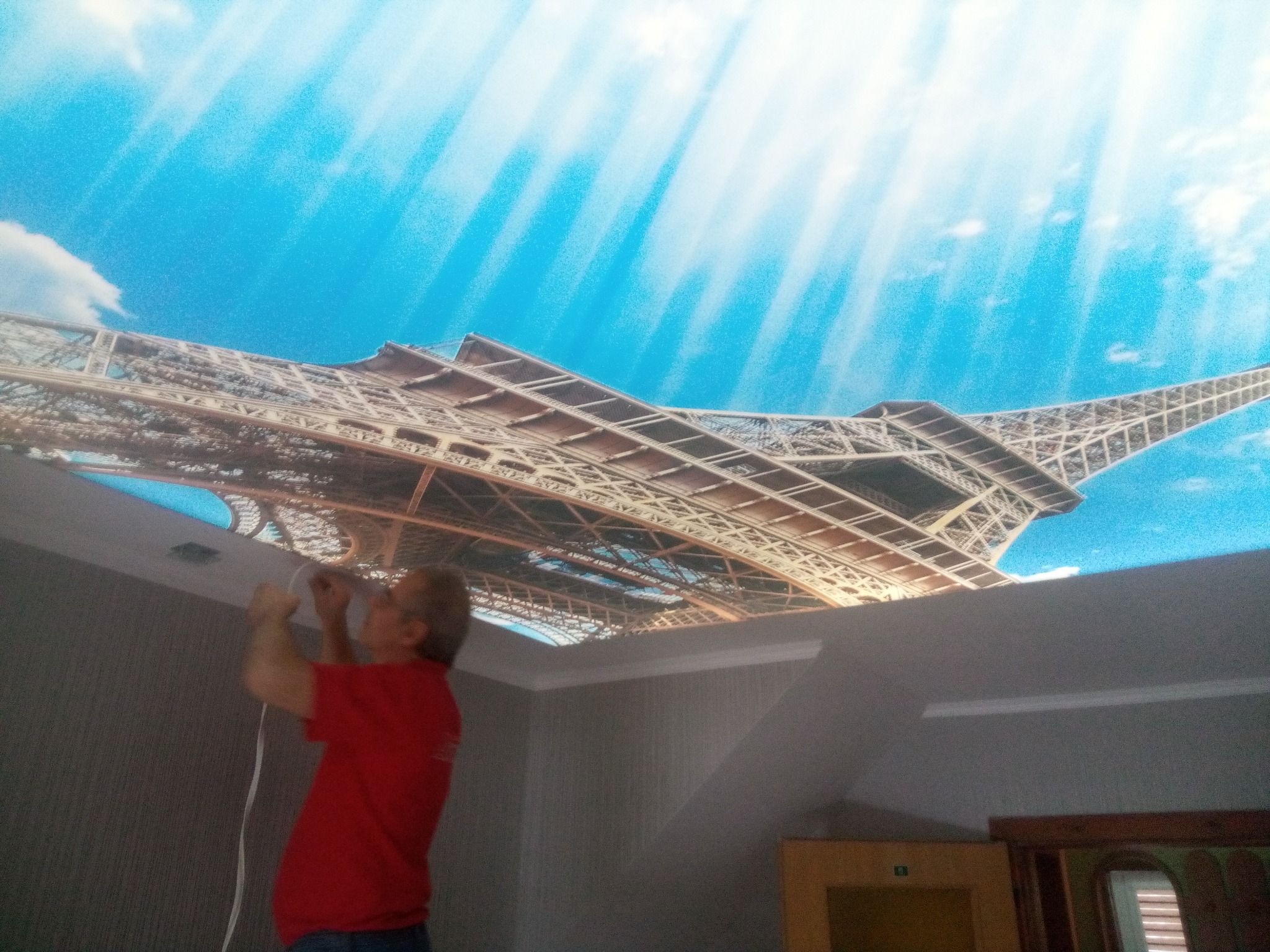 Montaż sufitu napinanego z motywem wieży Eiffla. Instalator w czerwonej koszulce mocuje konstrukcję. Widok z dołu na sufit i fragment ściany.