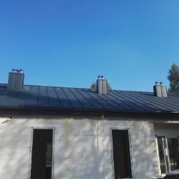 Nowy, grafitowy dach z blachy na surowym budynku z trzema kominami, błękitne niebo w tle.