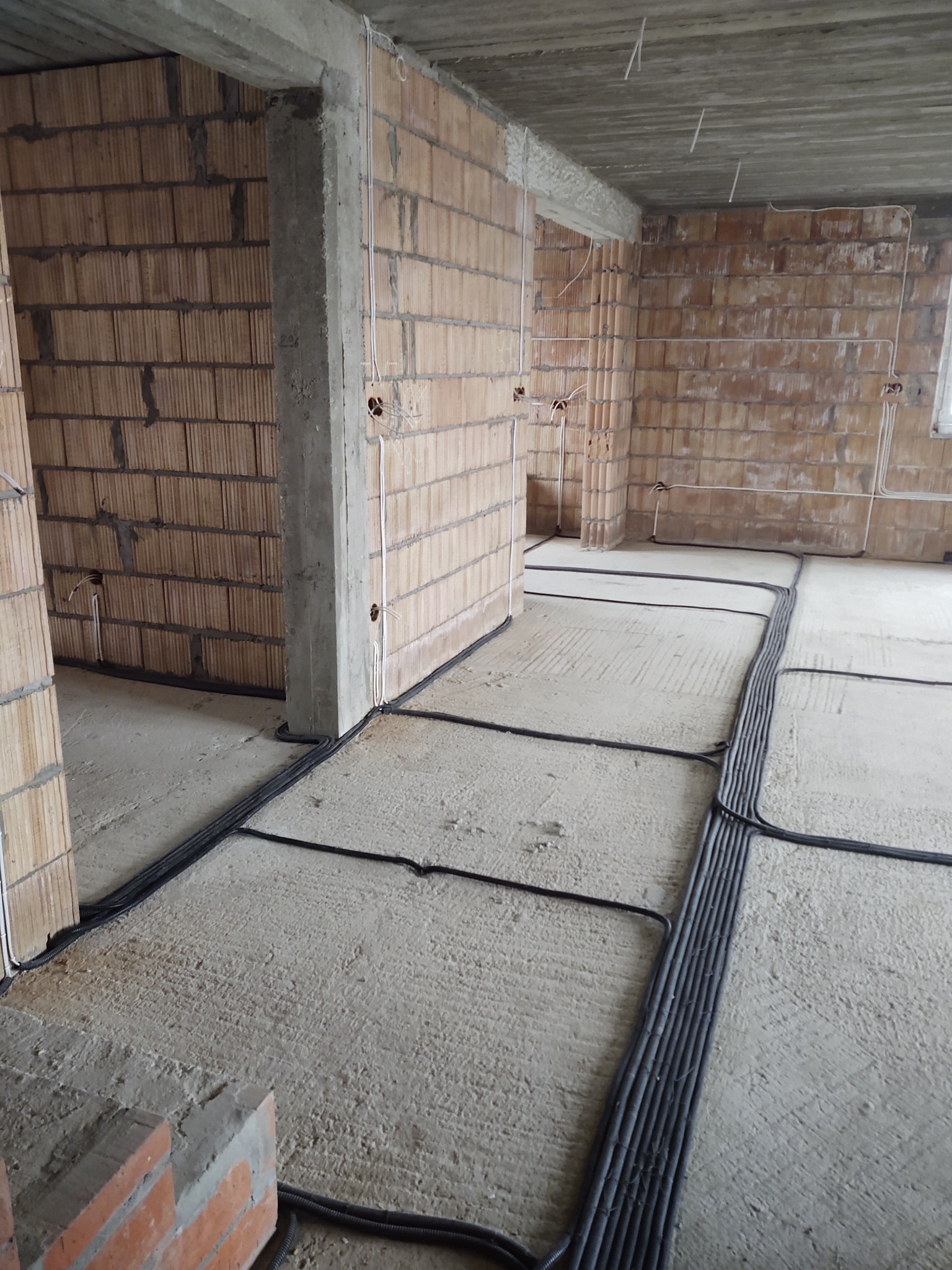 Surowy stan wnętrza budynku w trakcie prac instalacyjnych: widoczne przewody elektryczne ułożone na podłodze, prowadzone wzdłuż ścian z pustaków ceramicznych, wystające kable ze ścian i sufitu.