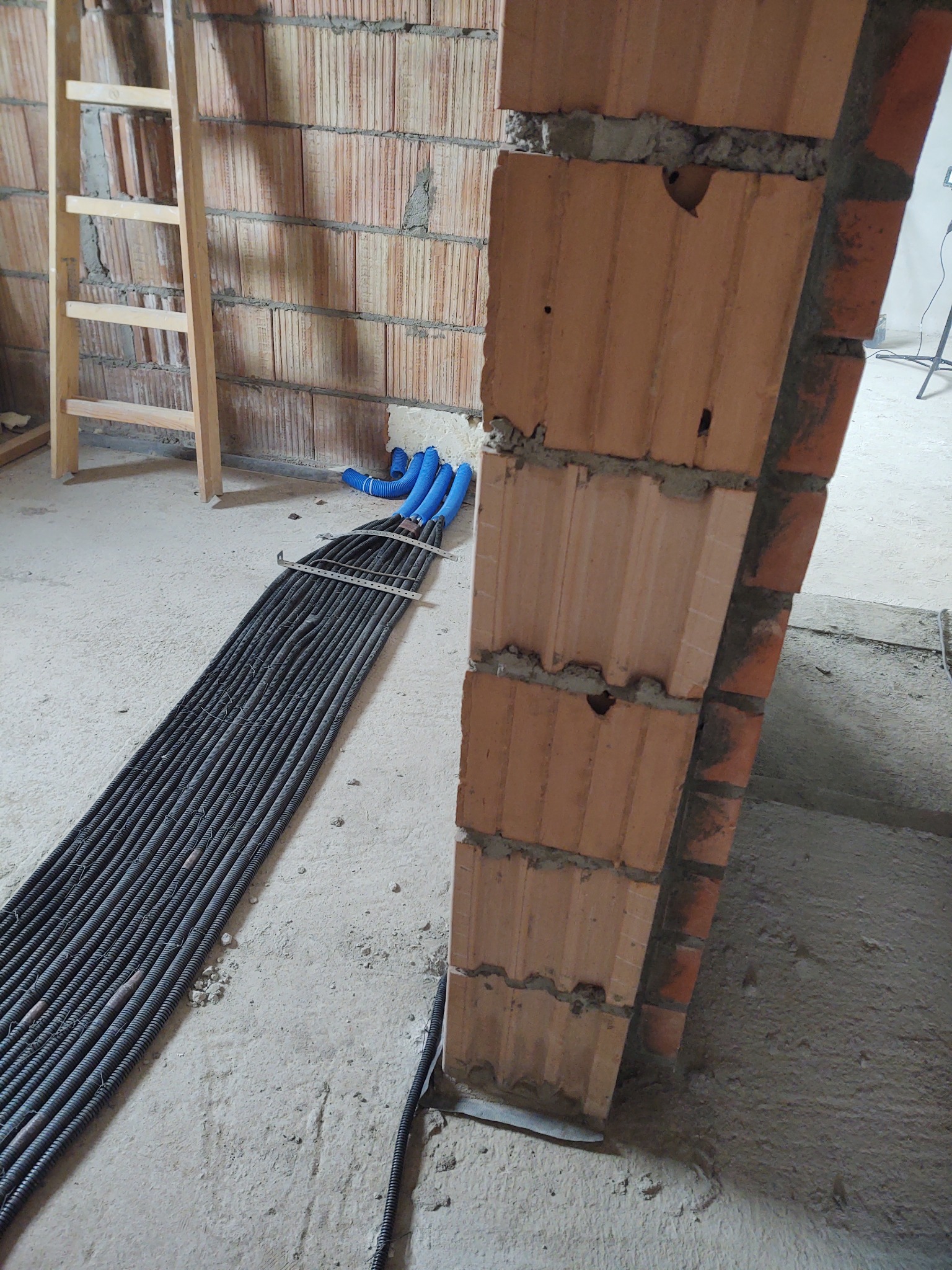 Wiązka przewodów elektrycznych w peszlach ułożona na surowej posadzce obok murowanego otworu drzwiowego z cegły kratówki.