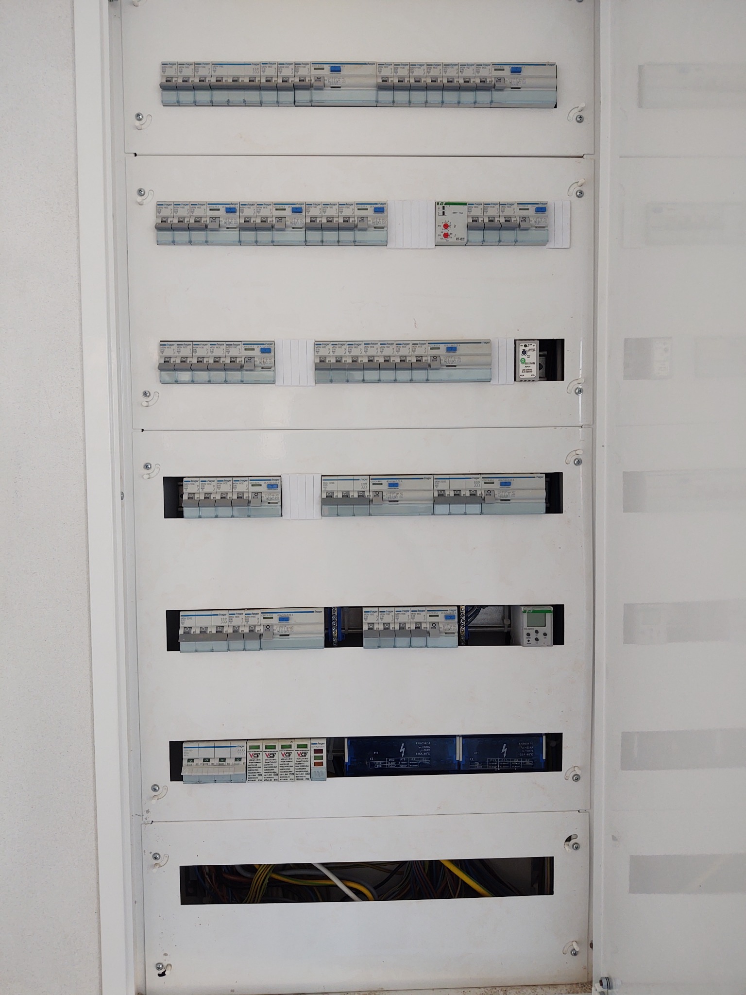 Otwarta, biała skrzynka rozdzielcza z widocznymi bezpiecznikami Hager, przekaźnikami i przewodami elektrycznymi w dolnej części.