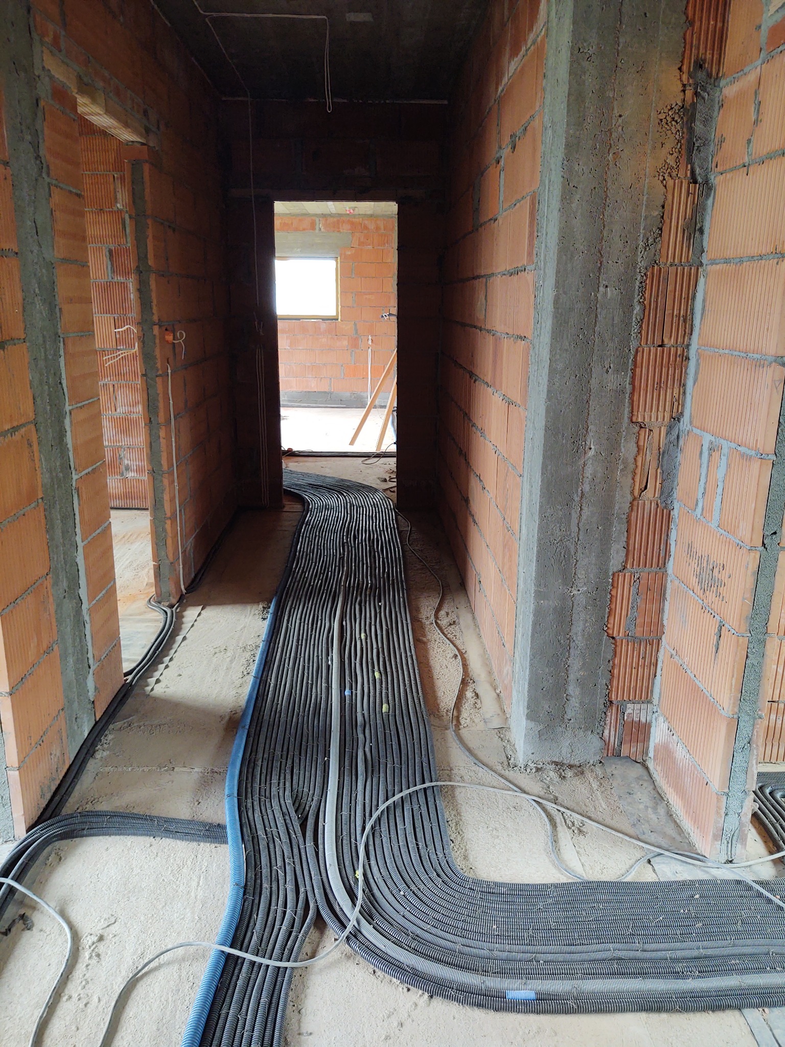 Wnętrze w trakcie budowy: liczne kable elektryczne ułożone na podłodze, prowadzące przez otwory w ścianach z cegły ceramicznej.