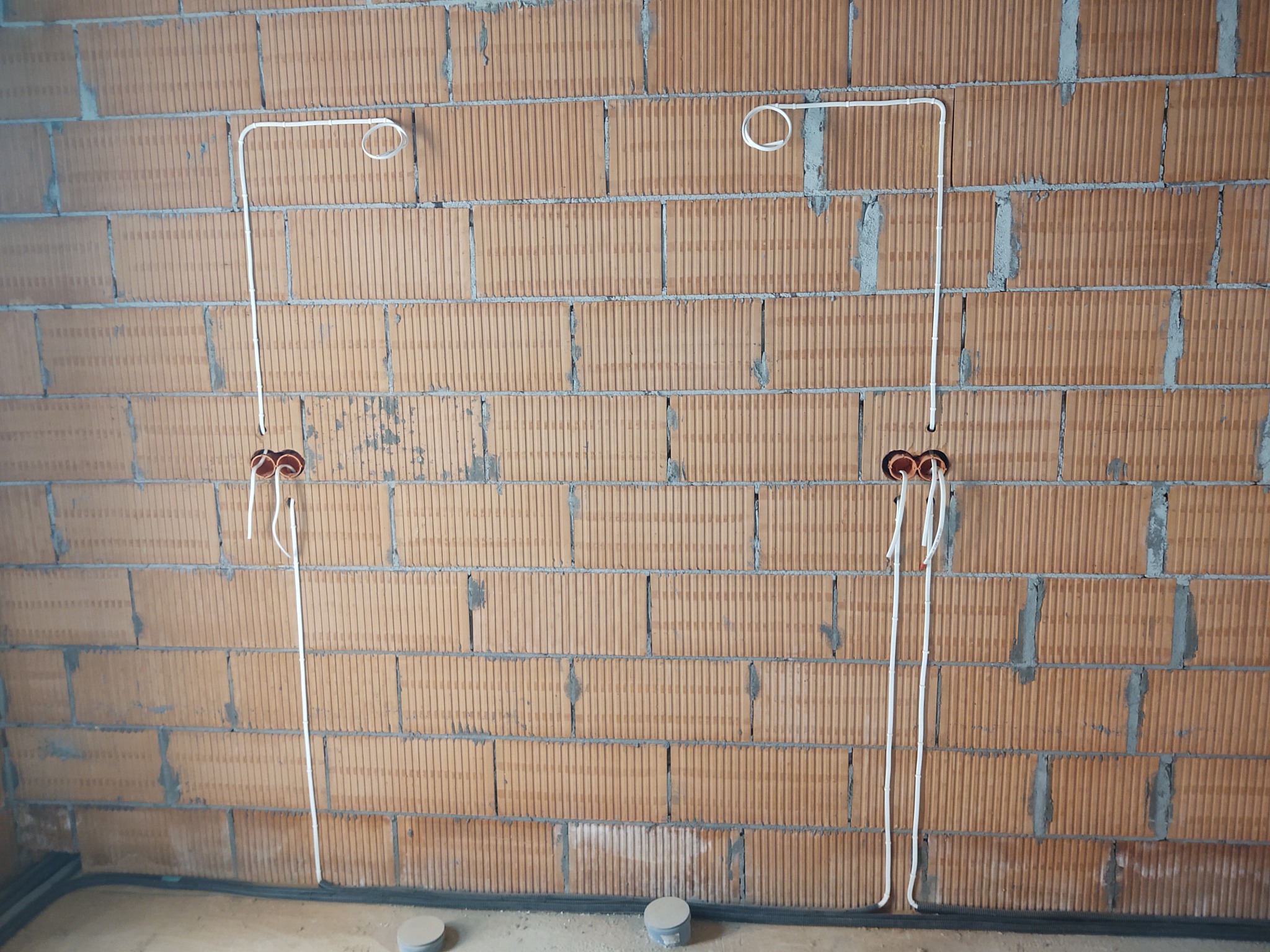 Białe przewody elektryczne ułożone na ceglanej ścianie z widocznymi puszkami instalacyjnymi i pętlami kablowymi pod sufitem.