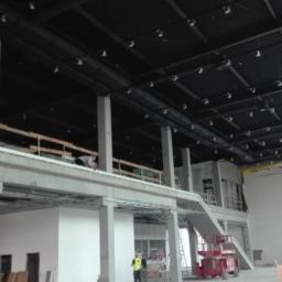 Mal-Nat - Wnętrze hali w stanie surowym, widoczne stalowe konstrukcje dachu z oświetleniem, surowe ściany, pracownicy w kamizelkach odblaskowych i podnośnik koszowy.