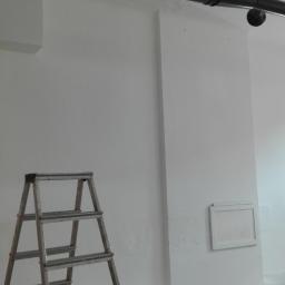 Mal-Nat - Wnętrze po świeżym malowaniu na biało, z widoczną metalową drabiną, rurami pod sufitem i otwartym przejściem do kolejnego pomieszczenia.