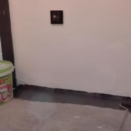 Mal-Nat - Wnętrze w trakcie odświeżania: wiadro z farbą 'Aksilbet' do betonu stoi obok świeżo pomalowanej na biało ściany, z widocznymi śladami wałka przy podłodze.