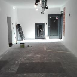 Mal-Nat - Przygotowane do malowania wnętrze biura z zabezpieczoną folią podłogą, widoczne wiadro z farbą, wałek i pędzel, surowe ściany i sufit przed malowaniem, industrialne lampy wiszące