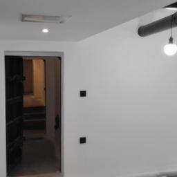 Mal-Nat - Świeżo pomalowane na biało wnętrze z widocznymi rurami pod sufitem i wiszącą żarówką, z otwartymi, ciemnymi drzwiami prowadzącymi do kolejnego pomieszczenia.