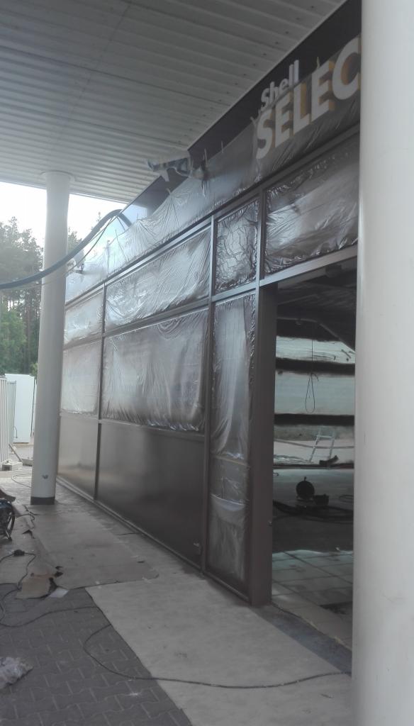 Przygotowanie do malowania elewacji budynku Shell Select, okna i elementy zabezpieczone folią ochronną, widoczne narzędzia malarskie.