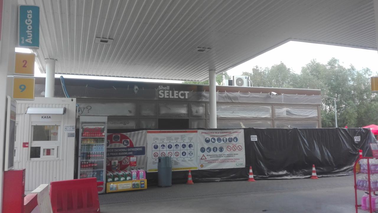Stacja benzynowa w trakcie prac remontowych: zabezpieczone okna folią, widoczne tablice informacyjne o pracach budowlanych, dystrybutor paliwa i kasa w tle.