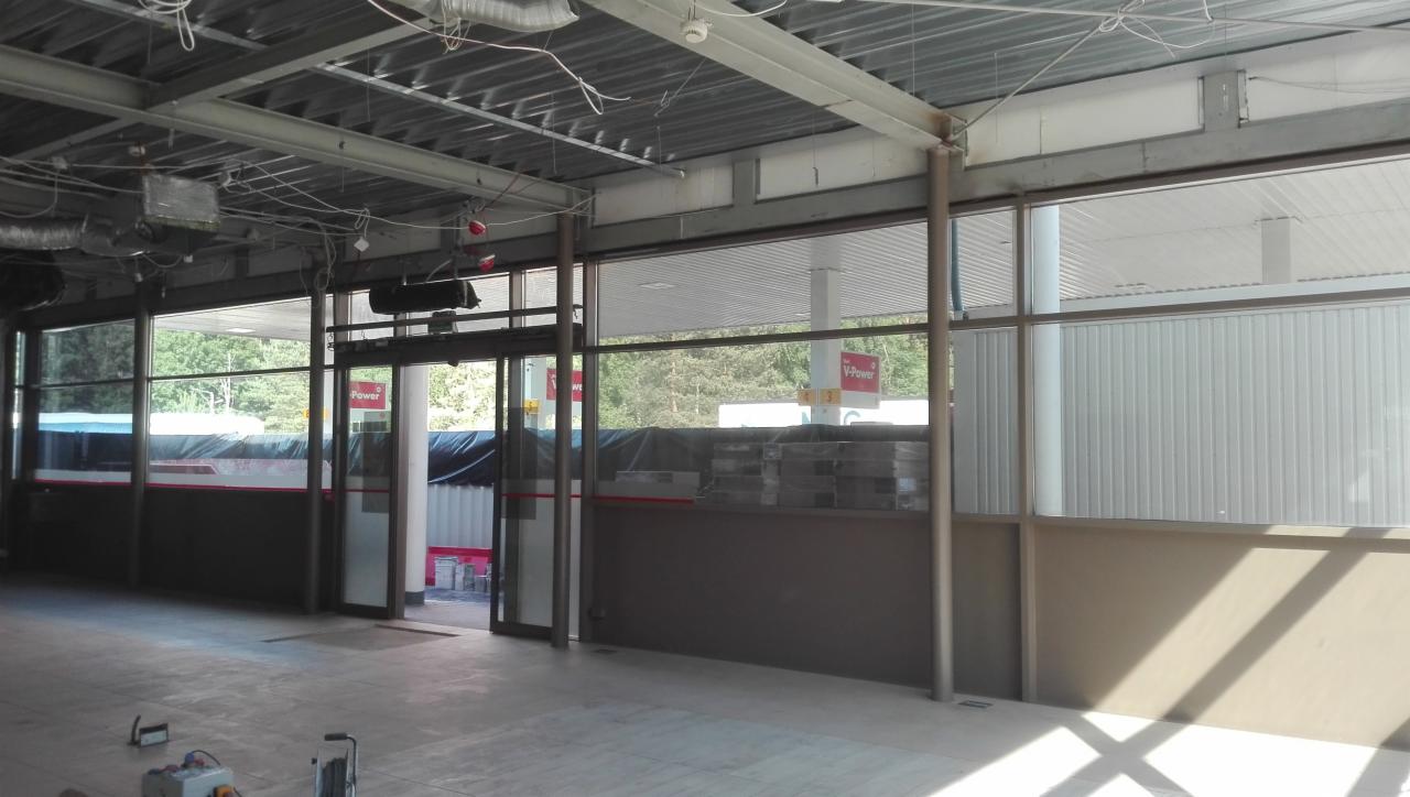 Wnętrze budynku w trakcie remontu z widocznymi elementami konstrukcyjnymi stropu, szklanymi ścianami i otwartymi drzwiami wejściowymi. Na podłodze widoczne narzędzia i materiały budowlane.