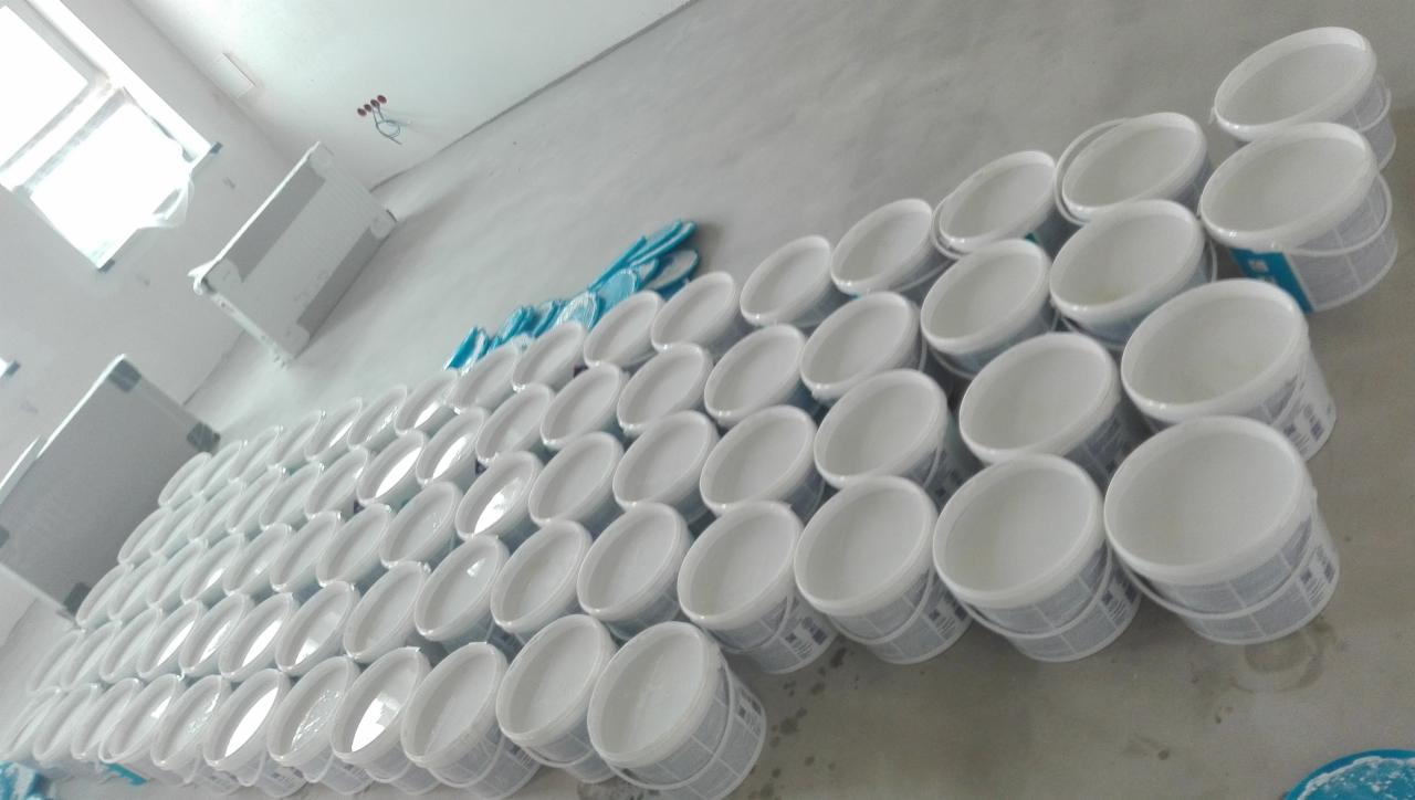 Rzędy białych, plastikowych wiader z farbą, częściowo przykrytych niebieską folią ochronną, ustawione na szarym, betonowym podłożu w pomieszczeniu przed malowaniem.