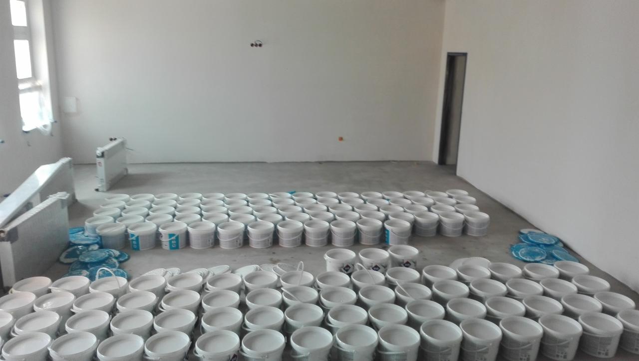 Przygotowanie do malowania: Rzędy białych wiader z farbą ustawione w pustym, jasnym pomieszczeniu z grzejnikami i zabezpieczonymi podłogami.