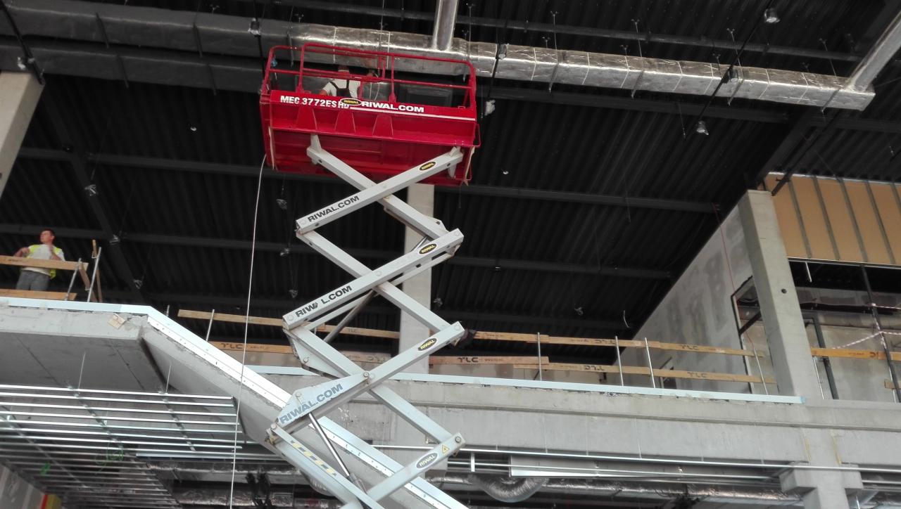 Wnętrze w trakcie prac wykończeniowych, widoczny podnośnik nożycowy z pracownikami na tle sufitu i instalacji wentylacyjnej.