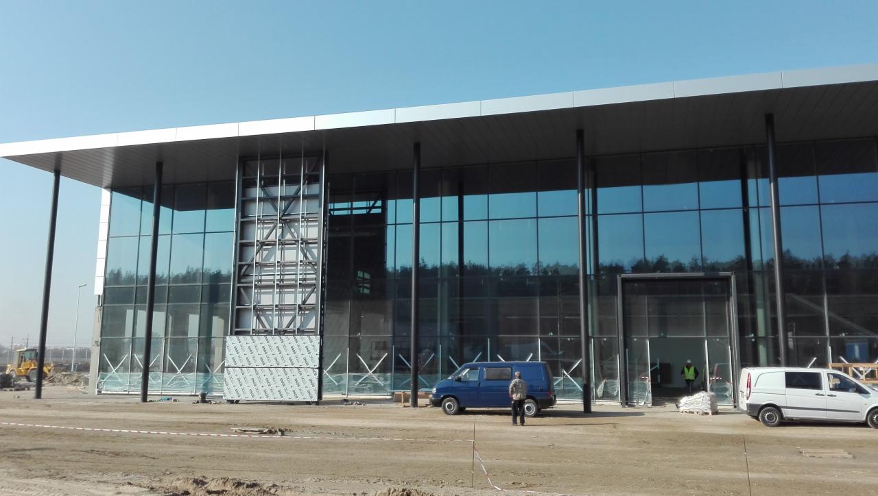 Budowa nowoczesnego budynku biurowego z przeszkloną fasadą i metalową konstrukcją, widoczne pojazdy budowlane i robotnicy.