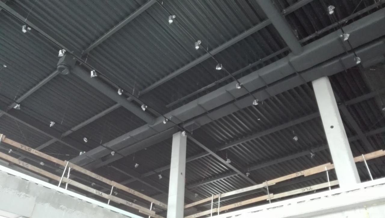 Metalowy sufit w trakcie prac wykończeniowych, widoczne instalacje wentylacyjne, oświetlenie punktowe i betonowe kolumny wsporcze.