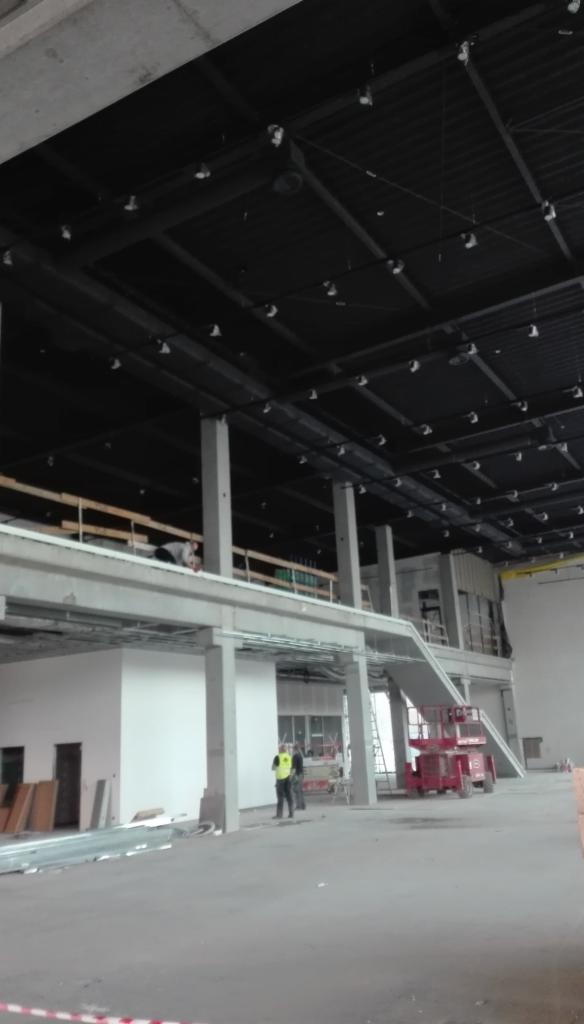 Wnętrze hali w stanie surowym, widoczne stalowe konstrukcje dachu z oświetleniem, surowe ściany, pracownicy w kamizelkach odblaskowych i podnośnik koszowy.