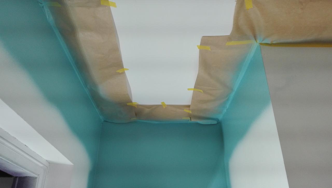 Świeżo pomalowany fragment pomieszczenia w odcieniach błękitu, zabezpieczony papierem maskującym i taśmą malarską na styku z sufitem i ścianą. Widoczny narożnik pokoju z fragmentem ościeżnicy.