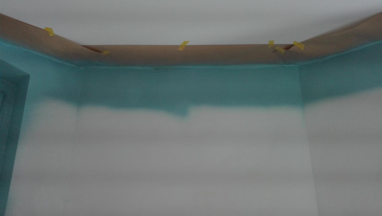 Ściana w trakcie malowania z częściowo nałożoną turkusową farbą, zabezpieczona taśmą malarską przy suficie i framudze drzwi.