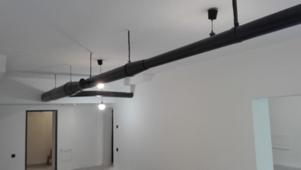 Czarny system rur wentylacyjnych pod sufitem w świeżo wykończonym, białym pomieszczeniu z widocznymi lampami wiszącymi i otworami drzwiowymi.