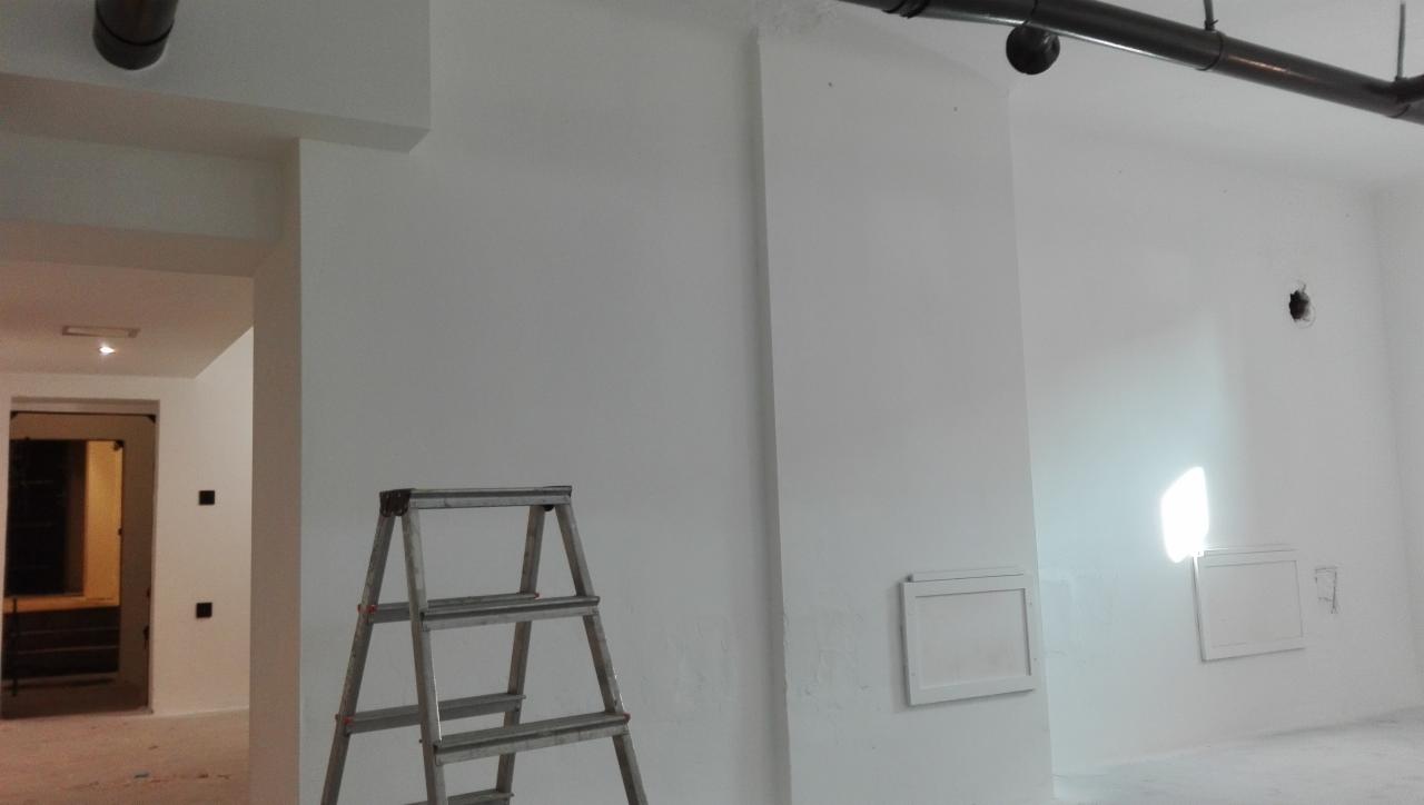 Wnętrze po świeżym malowaniu na biało, z widoczną metalową drabiną, rurami pod sufitem i otwartym przejściem do kolejnego pomieszczenia.