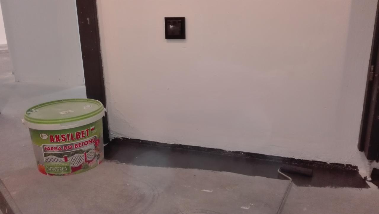 Wnętrze w trakcie odświeżania: wiadro z farbą 'Aksilbet' do betonu stoi obok świeżo pomalowanej na biało ściany, z widocznymi śladami wałka przy podłodze.
