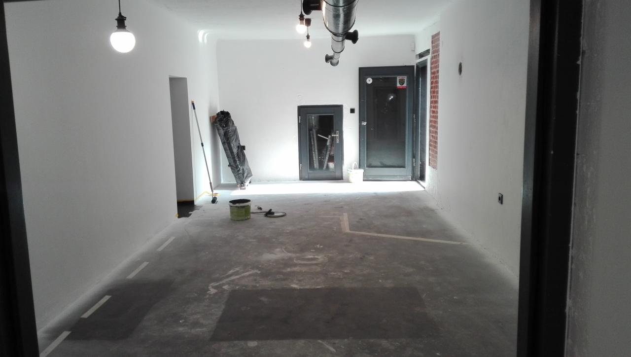 Przygotowane do malowania wnętrze biura z zabezpieczoną folią podłogą, widoczne wiadro z farbą, wałek i pędzel, surowe ściany i sufit przed malowaniem, industrialne lampy wiszące