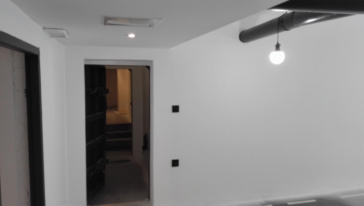 Świeżo pomalowane na biało wnętrze z widocznymi rurami pod sufitem i wiszącą żarówką, z otwartymi, ciemnymi drzwiami prowadzącymi do kolejnego pomieszczenia.