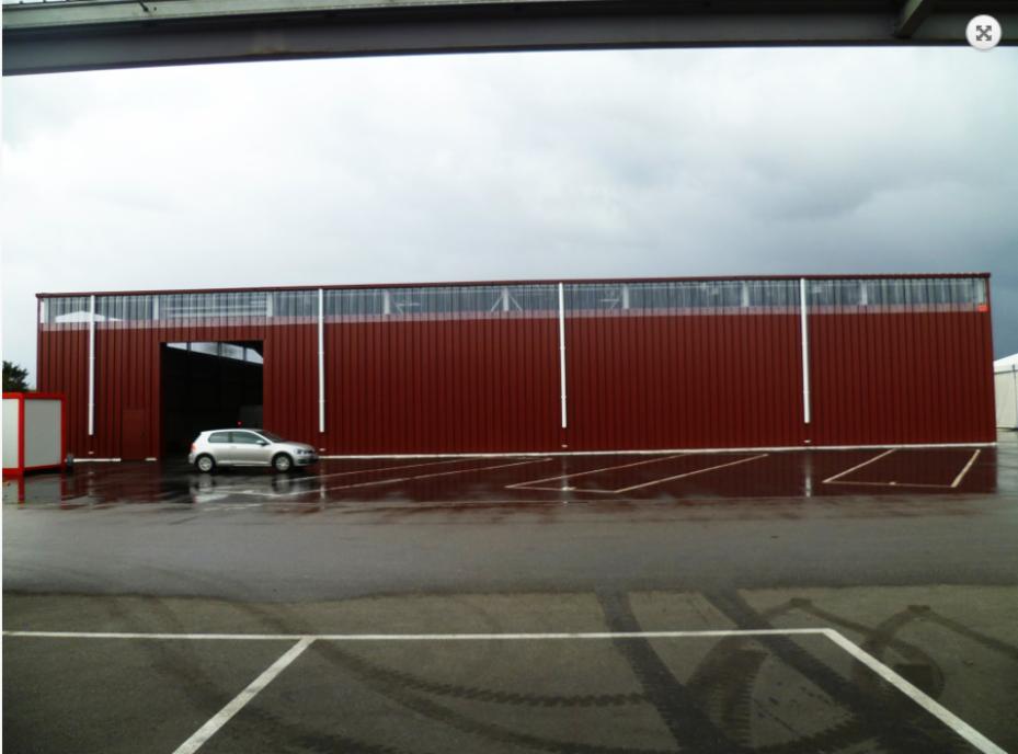 Duży, bordowy budynek przemysłowy z blachy falistej, z otwartą bramą wjazdową, z widocznym srebrnym samochodem osobowym na mokrym parkingu w pochmurny dzień.