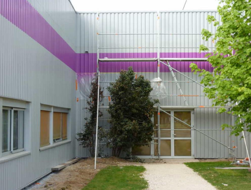 Elewacja budynku biurowego z szarej blachy trapezowej z fioletowym pasem, w trakcie prac z użyciem rusztowania, zabezpieczona folią malarską.